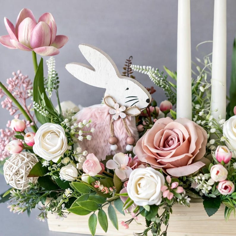 May include: An Easter floral arrangement featuring a white wooden rabbit with a pink fluffy body, set amongst pink and white flowers, greenery, and two white candles in a wooden box. The display includes roses, tulips, and other spring flowers.