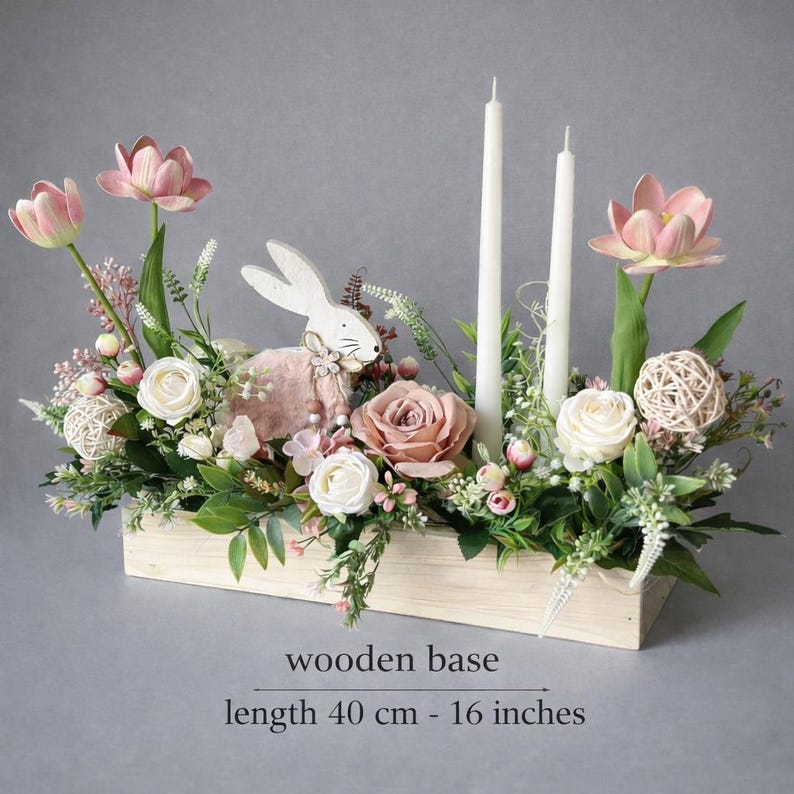 May include: An Easter centrepiece in a wooden base, 40 cm long. It features pink and white flowers, greenery, two white candles, and a wooden bunny figurine. The arrangement includes roses, tulips, and decorative spheres.