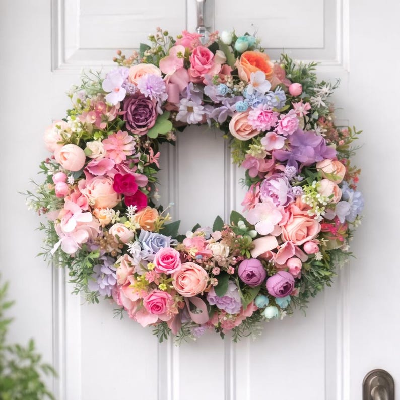 May include: A vibrant floral wreath featuring an array of flowers in pink, purple, peach, and blue hues. The circular wreath is composed of artificial flowers and foliage, suitable for spring or summer decoration.