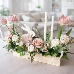 May include: A wooden box arrangement featuring pink and white flowers, greenery, and two white candles. A decorative white bunny and a pink lamb are also included. The arrangement is set on a light-coloured surface.
