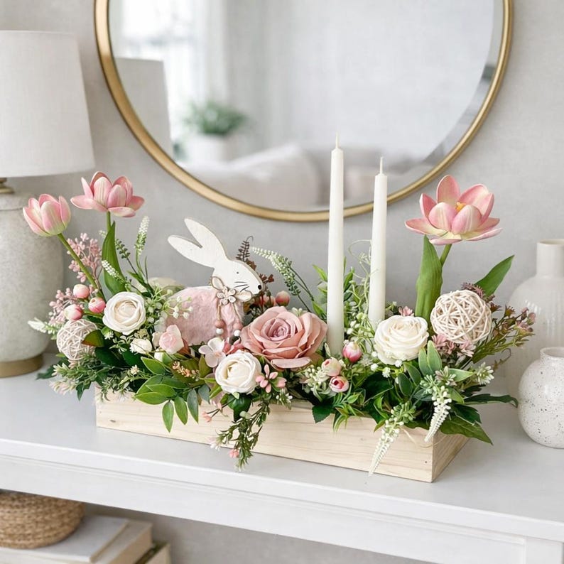May include: A wooden rectangular floral arrangement featuring pink and white flowers, greenery, and two white candles. A white bunny figurine with a pink body is in the centre. The arrangement is on a white surface, with a gold-framed mirror behind.