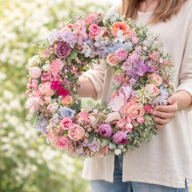 May include: A colourful floral wreath composed of roses, peonies, and other flowers in pastel shades. The circular wreath, with pink, purple, and peach blossoms, is held in front of a blurred background of greenery.