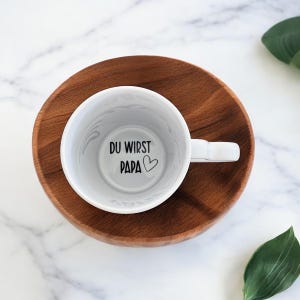 Könnte beinhalten: Eine weiße Keramik-Tasse mit dem Text "DU WIRST PAPA" und einem Herzsymbol im Inneren. Die Tasse steht auf einem runden Holztablett. Der Hintergrund ist eine weiße Marmoroberfläche mit einem grünen Blatt.