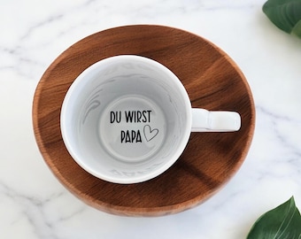 Puede incluir: Una taza de cerámica blanca con el texto "DU WIRST PAPA" y un símbolo de corazón en su interior. La taza está sobre un plato redondo de madera. El fondo es una superficie de mármol blanco con una hoja verde.