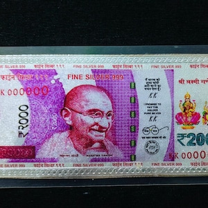 Puede incluir: Un billete de banco ₹2000 enmarcado en plata, morado y blanco, con Mahatma Gandhi e imágenes de deidades. El billete está marcado "FINE SILVER 999" e incluye el texto "I promise to pay the holder pure silver."