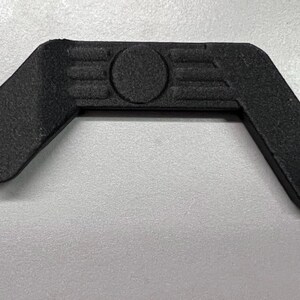 May include: A black, angular component with a textured surface. It features a rounded lever, a central circular detail, and a right-angled section with two holes. The object is likely a part of a larger mechanism.