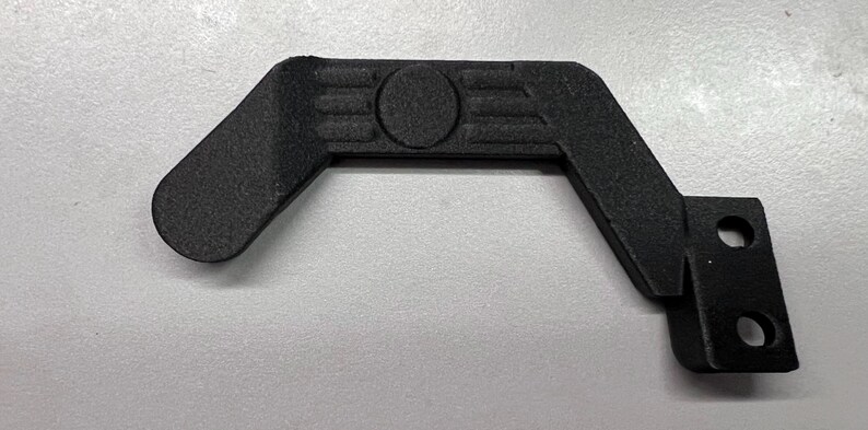 May include: A black, angular component with a textured surface. It features a rounded lever, a central circular detail, and a right-angled section with two holes. The object is likely a part of a larger mechanism.