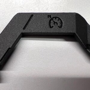 May include: A black, angular component with a textured surface. It features two circular holes, a diagonal line, and a circular emblem with a stylized design. The component has a bent shape with a series of parallel lines at one end.