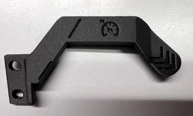 May include: A black, angular component with a textured surface. It features two circular holes, a diagonal line, and a circular emblem with a stylized design. The component has a bent shape with a series of parallel lines at one end.
