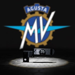 May include: A black motorcycle component with a circular gauge and a small, rectangular section. The blurred MV Agusta logo in white, blue, and gold is in the background against a dark backdrop.
