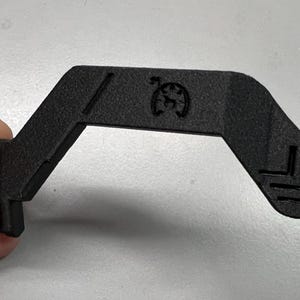 May include: A black, angular plastic component with a distinctive shape. It features a small circular emblem and angled lines. The component is held by a hand, showing its size and design. The object has a textured surface.