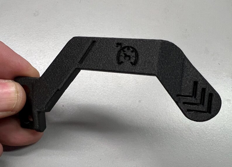 May include: A black, angular plastic component with a distinctive shape. It features a small circular emblem and angled lines. The component is held by a hand, showing its size and design. The object has a textured surface.