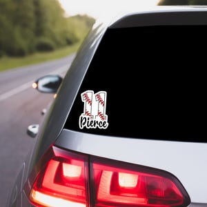 May include: A white car window sticker with the number "11" designed as baseballs, and the name "Pierce" in a script font. The sticker is on the rear window of a silver car, with a road and trees in the background.