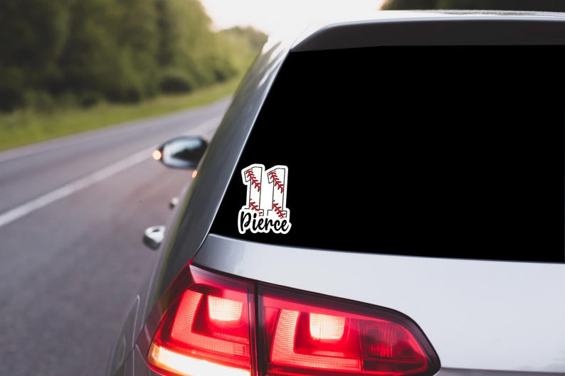 May include: A white car window sticker with the number "11" designed as baseballs, and the name "Pierce" in a script font. The sticker is on the rear window of a silver car, with a road and trees in the background.
