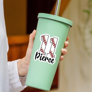 May include: A light green tumbler with a silver straw. A white sticker with the number "11" in a baseball design and the name "Pierce" in a script font is on the tumbler. The tumbler is held by a person.