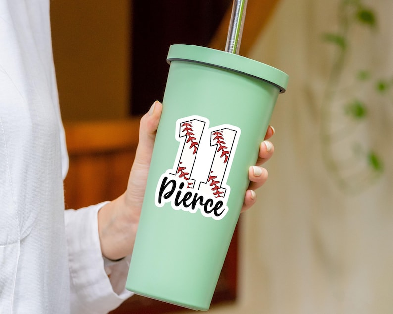 May include: A light green tumbler with a silver straw. A white sticker with the number "11" in a baseball design and the name "Pierce" in a script font is on the tumbler. The tumbler is held by a person.