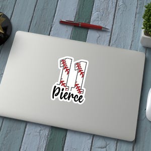 May include: A white sticker on a laptop with the number "11" in a baseball-style design, with red stitching and the name "Pierce" in a script font. The laptop is on a blue wooden surface, with a pen, mouse, and decorative items.