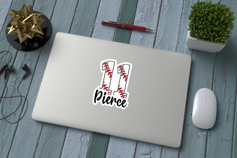 May include: A white sticker on a laptop with the number "11" in a baseball-style design, with red stitching and the name "Pierce" in a script font. The laptop is on a blue wooden surface, with a pen, mouse, and decorative items.