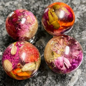 Op de afbeelding: Vier heldere, bolvormige harsobjecten, elk met gedroogde bloemen in tinten roze, oranje en geel. De bollen hebben een glanzende afwerking en staan op een donkergrijze ondergrond. De bloemen zijn zichtbaar in de transparante hars.