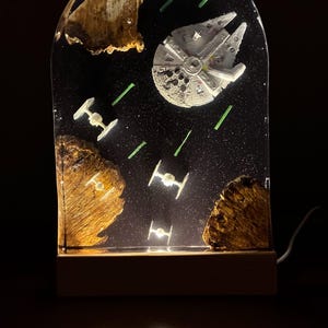 Könnte beinhalten: Eine Star Wars-Harzlampe mit dem Millennium Falken und TIE-Jägern vor einem sternenklaren schwarzen Hintergrund. Die Lampe enthält grüne Laserfeuer und zwei braune Felsformationen. Der Sockel besteht aus hellem Holz.