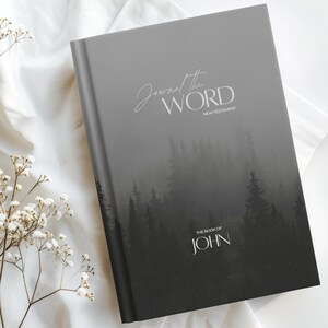 Könnte beinhalten: Ein Hardcover-Journal mit dem Text "Journal the Word" und "New Testament" in weißer Schrift. Das Cover zeigt eine Graustufen-Waldszene und die Worte "The Book of John". Das Journal liegt auf einer weißen Satin-Oberfläche.