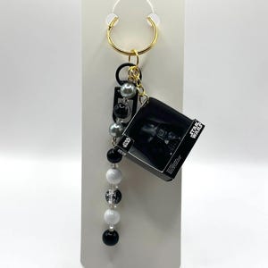 May include: A Star Wars themed keychain featuring a small box with a Darth Vader image. The keychain has a gold-colored ring, black and silver beads, and a dangling bead design with black, white, and clear beads.