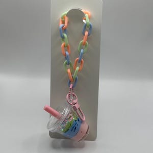 May include: A pastel-colored keychain featuring a miniature bubble tea cup charm. The cup is clear with pink, blue, and green accents, and a pink straw. The keychain has a chain of pastel orange, green, and blue links, and a pink clasp.