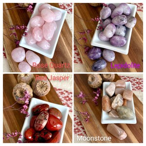 May include: Four white rectangular dishes filled with polished gemstones: rose quartz, lepidolite, red jasper, and moonstone. Each dish is on a wooden board with dried flowers. Text labels identify each gemstone type.