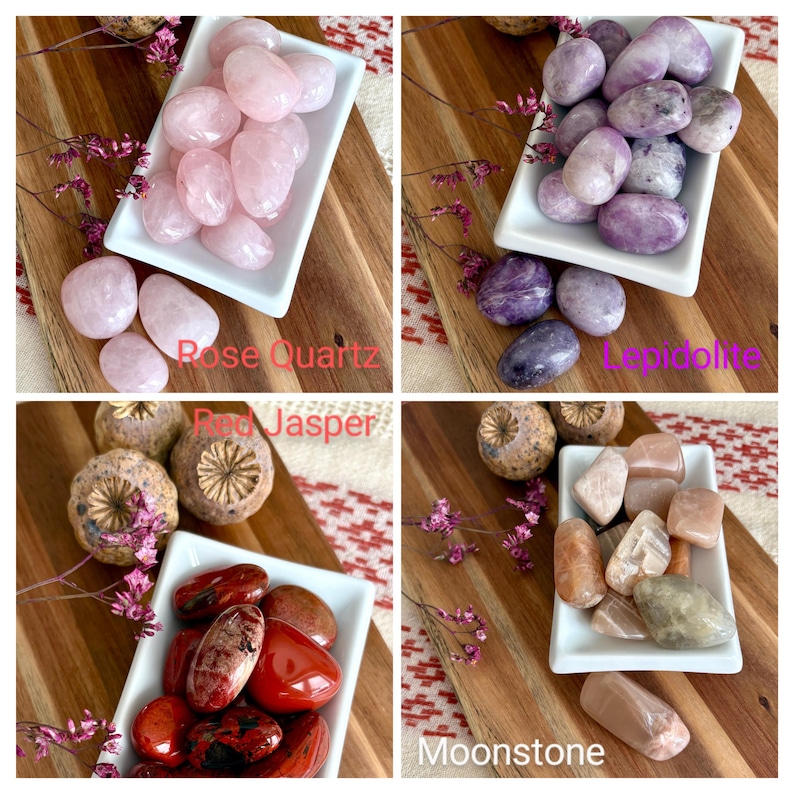 May include: Four white rectangular dishes filled with polished gemstones: rose quartz, lepidolite, red jasper, and moonstone. Each dish is on a wooden board with dried flowers. Text labels identify each gemstone type.