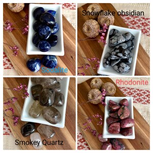 May include: A collection of polished gemstones, including sodalite, snowflake obsidian, smokey quartz, and rhodonite, displayed in white rectangular dishes on a wooden board. The image includes text labels for each stone type and decorative dried flowers.