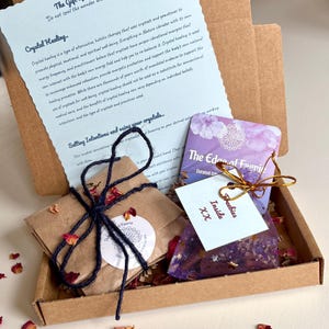 May include: A gift box containing a small package tied with navy twine and dried rose petals, a purple sachet, and a card that reads "Inside Freebies XX". A printed card with text about crystal healing is also included.