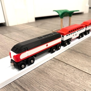 Può includere: Un set di trenini giocattolo in legno con una locomotiva nera e rossa e vagoni passeggeri abbinati. Il treno è su un binario bianco, con una stazione verde e beige sullo sfondo. I vagoni hanno il tetto rosso e i lati bianchi.