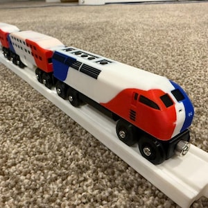 Può includere: Un set di trenini giocattolo in legno con un binario bianco. I vagoni sono rossi, bianchi e blu. La locomotiva ha un frontale rosso e blu. Il treno è su un binario bianco.