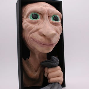 May include: A three-dimensional wall sculpture of a pale, large-eared creature with prominent green eyes and wrinkled skin, set within a rectangular black frame. The figure is depicted from the chest up, holding a dark gray fabric item in one hand.