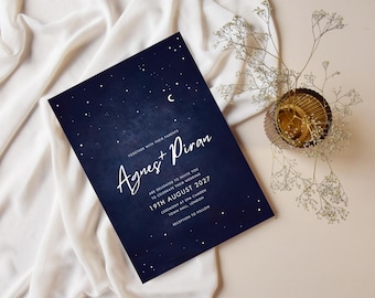 May include: A navy blue wedding invitation with white script that reads "Agnes + Piran" and the date "19th August 2027". The invitation has a starry night design and is placed on a white cloth with a small gold-colored decorative bowl.