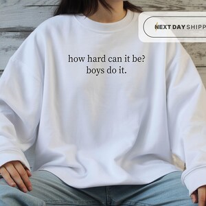 May include: A white long-sleeve crewneck sweatshirt featuring the black printed text how hard can it be? boys do it. centered on the chest. The garment has a ribbed neckline, cuffs, and hem, and is worn with light blue denim jeans.