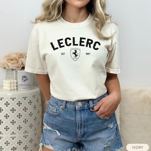 May include: Ivory-colored t-shirt featuring the name "LECLERC" in bold black letters above a shield with a Ferrari logo and the year "1997." The shirt is a casual style, suitable for everyday wear.