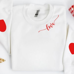 May include: White sweatshirt with red heart graphics on the sleeves and the word "love" in red script on the chest. The sweatshirt is a crew neck style and is likely made of a soft material. A festive gift box is in the background.