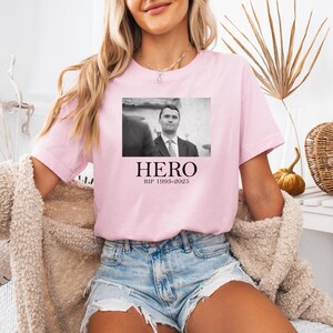 A woman wearing a pink t-shirt with a black and white photo of a man and the text "HERO RIP 1968-2005" printed on it.