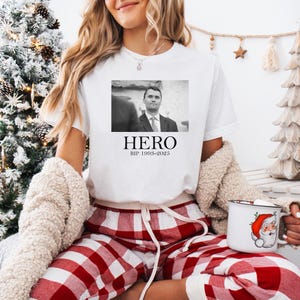 A woman wearing a white t-shirt with a black and white photo of a man and the text "HERO" is sitting in front of a Christmas tree. She is holding a mug with a Santa Claus design.