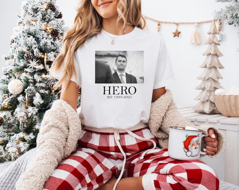 A woman wearing a white t-shirt with a black and white photo of a man and the text "HERO" is sitting in front of a Christmas tree. She is holding a mug with a Santa Claus design.