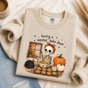 May include: A beige crewneck sweatshirt featuring a graphic of a skeleton wearing a polka dot headscarf and apron, baking cookies in a kitchen. The text reads having a mental bake down, accompanied by a black cat, pumpkins, and a window.