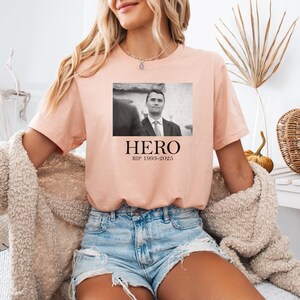 A woman wearing a pink t-shirt with a black and white photo of a man and the text "HERO Rip 1989-2025" printed on it, sitting in a chair with a beige sweater and denim shorts.