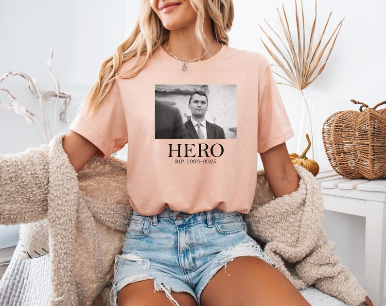 A woman wearing a pink t-shirt with a black and white photo of a man and the text "HERO Rip 1989-2025" printed on it, sitting in a chair with a beige sweater and denim shorts.