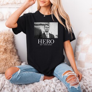 A woman wearing a black t-shirt with a black and white photo of a man and the text "HERO" and "RIP 1995-2025" printed on it, sitting on a couch.