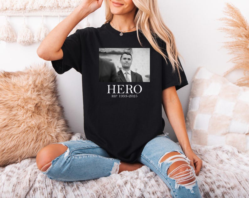 A woman wearing a black t-shirt with a black and white photo of a man and the text "HERO" and "RIP 1995-2025" printed on it, sitting on a couch.