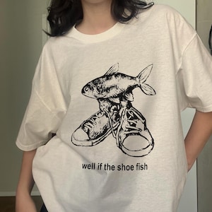 May include: White t-shirt with a black graphic of a fish perched atop a pair of sneakers. The text "well if the shoe fish" is printed below the graphic. The shirt has short sleeves and a crew neck.