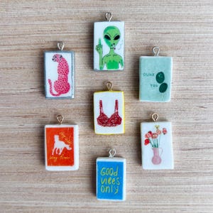 May include: A collection of rectangular pendants with various designs. One features a pink cheetah, another an alien smoking and giving the middle finger. Other pendants display a red bra, flowers in a vase, and the text "Good Vibes Only."