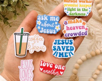 May include: A collection of seven vinyl stickers featuring religious phrases and graphics. Designs include a coffee cup, text reading ask me about Jesus, waymaker miracle worker, make heaven crowded, Jesus saved me, coffee & Jesus, Jesus loves you, and love one another.