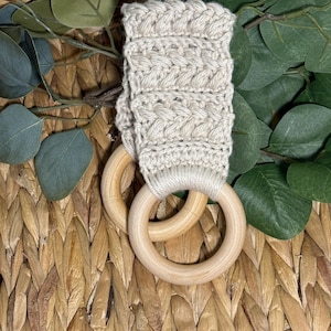 Könnte beinhalten: Ein beige gehäkelter Beißring für Babys mit zwei Holzringen. Der Beißring besteht aus einem weichen, strukturierten Garn und ist an den Holzringen befestigt. Der Hintergrund ist ein geflochtener Korb mit grünen Blättern.